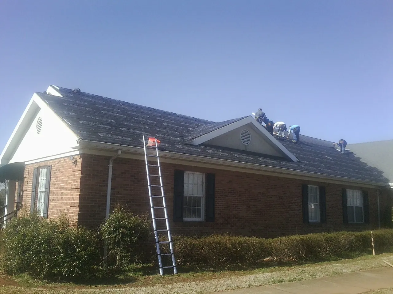 Crew tearing shingles off a brick ranch roof with a ladder set — Emergency Roof Repair work by our crew in White Stone