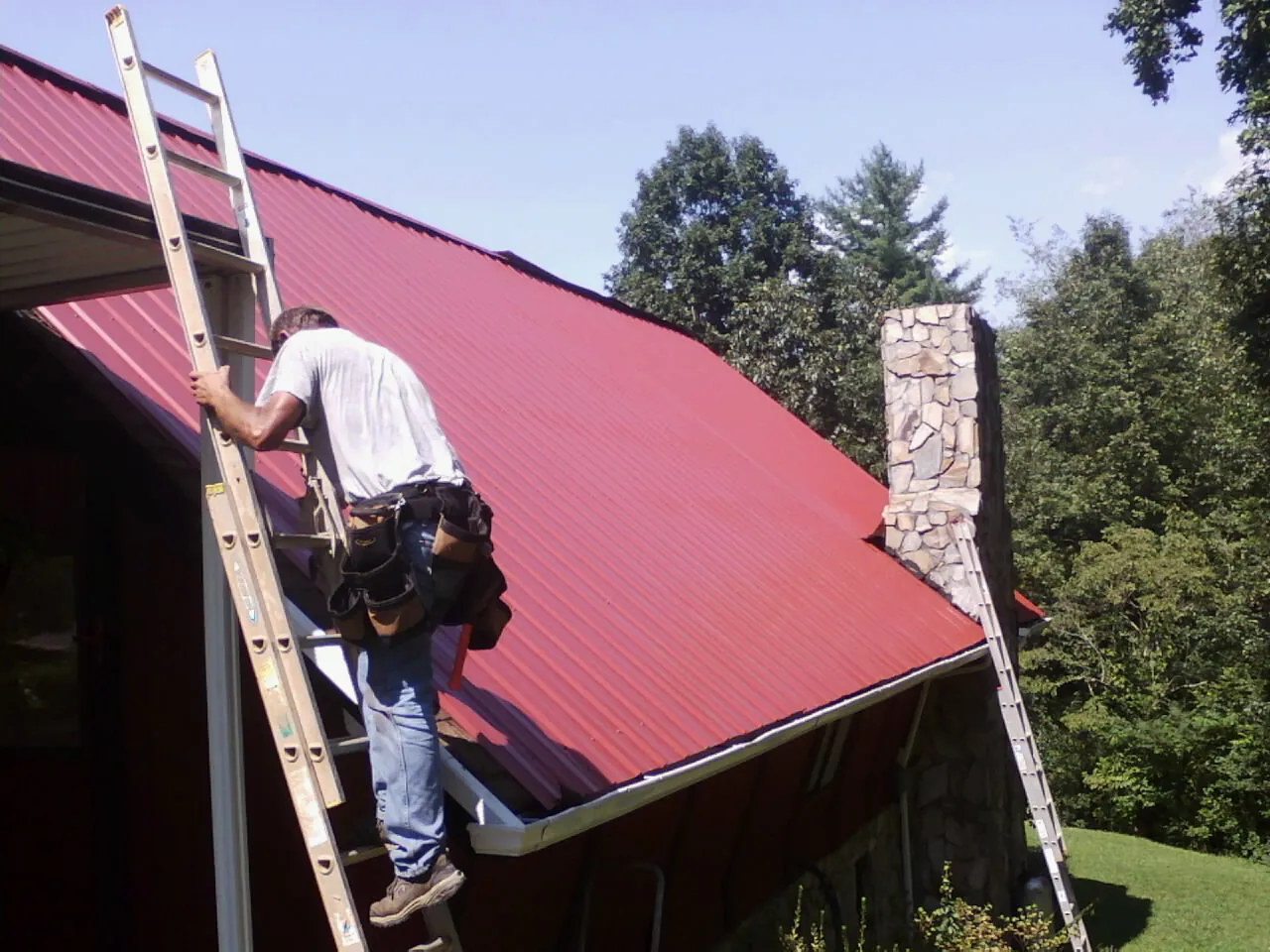 Roofer on a ladder beside a red metal roof and stone chimney — Soffit & Fascia Repair by Roofing Brothers in White Stone